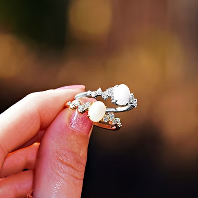 A close-up of a hand holding two silver rings with oval white stones and small clear accent stones, set against a warm, blurred bokeh background.