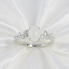 A silver ring featuring a large oval white stone in the center, flanked by small clear stones on both sides, displayed on a white satin fabric background.