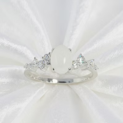 A silver ring featuring a large oval white stone in the center, flanked by small clear stones on both sides, displayed on a white satin fabric background.
