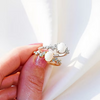 A close-up of a hand wearing two delicate rings with white oval stones and small clear crystal accents, set against a bright white background with soft shadows.