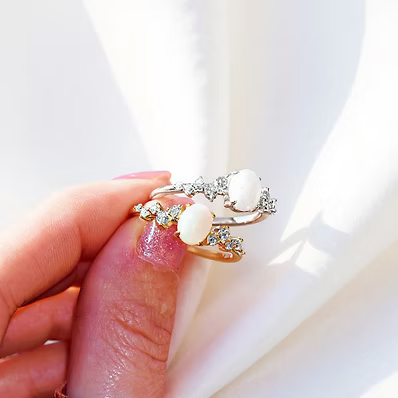 A close-up of a hand wearing two delicate rings with white oval stones and small clear crystal accents, set against a bright white background with soft shadows.