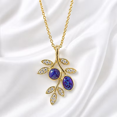 A gold leaf-shaped pendant necklace featuring two oval blue opal gemstones and small clear crystal accents, displayed on a white satin fabric background.