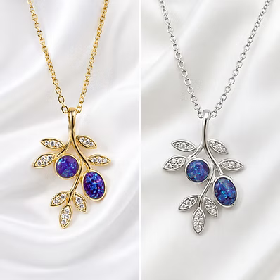 Two leaf-shaped pendant necklaces displayed side by side on a white satin background. The left necklace is gold-toned with two oval blue opal stones and small clear crystal accents.