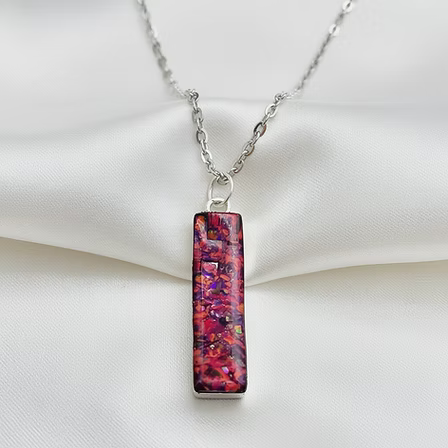 A silver chain necklace with a rectangular pendant featuring a vibrant, opal pattern of pink, red, purple, and black, displayed on a draped white satin fabric.