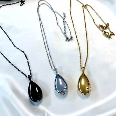Three teardrop pendant necklaces displayed on a white satin-like fabric background, shown in black, silver, and gold finishes.