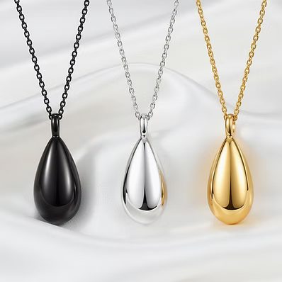 Three teardrop-shaped pendant necklaces displayed side by side on a white satin fabric background, shown in black, silver, and gold finishes.