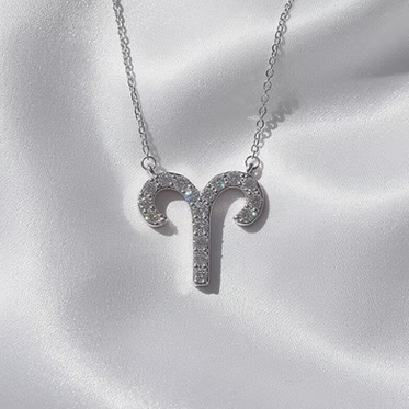 A silver-toned Aries zodiac sign pendant necklace adorned with small clear crystals, displayed on a soft white satin fabric background.
