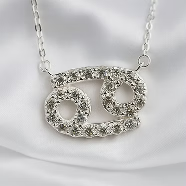A silver-tone necklace with a Gemini zodiac symbol pendant, embellished with clear rhinestones, displayed on a white satin fabric background.