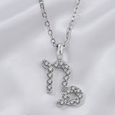 A silver-tone pendant necklace featuring the Capricorn zodiac symbol, embellished with small clear stones, displayed on a white satin fabric background.