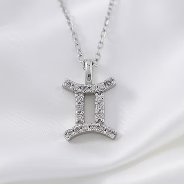 A silver-tone Gemini zodiac sign pendant necklace adorned with small clear crystals, resting on a white satin fabric background.