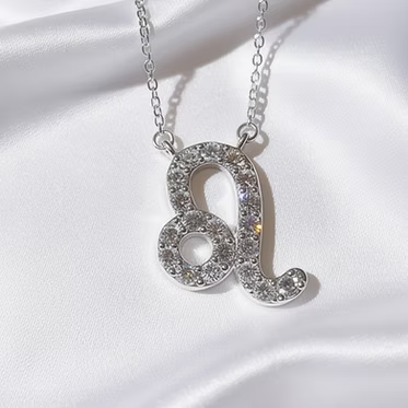 A silver-tone necklace with a Leo zodiac symbol pendant, set with sparkling clear crystals, is displayed on draped white satin.