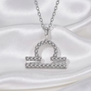 A silver-tone necklace with a Libra zodiac sign pendant, embellished with small clear crystals, displayed on a draped white satin fabric.