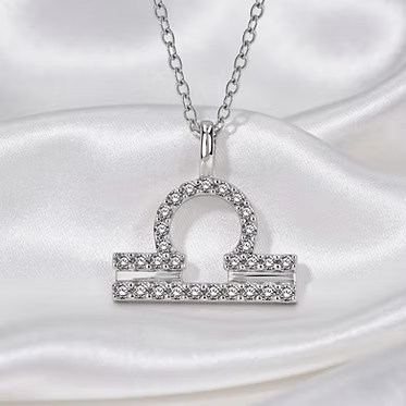 A silver-tone necklace with a Libra zodiac sign pendant, embellished with small clear crystals, displayed on a draped white satin fabric.