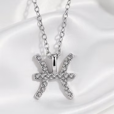 A silver-toned Pisces zodiac sign pendant necklace adorned with small clear crystals, displayed on a white satin fabric background.