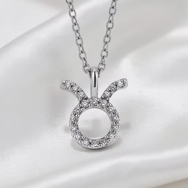 A silver-toned necklace with a Zodiac Aries pendant, featuring a halo of small clear stones around an open circle with curved horns, displayed on a white satin fabric background.