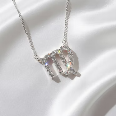 A silver-toned necklace with a Virgo zodiac sign pendant, embellished with sparkling clear crystals, is displayed on a draped white satin fabric.