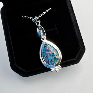 A teardrop-shaped pendant necklace featuring a vibrant blue, green, and orange iridescent opal stone in a polished silver bezel, displayed on a black velvet jewelry box.