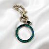 A silver-tone keychain or bag charm featuring a large open circular ring inlaid with iridescent green and blue abalone shell, a small dark green teardrop gemstone, and a heart-shaped clasp, displayed on a white satin fabric background.
