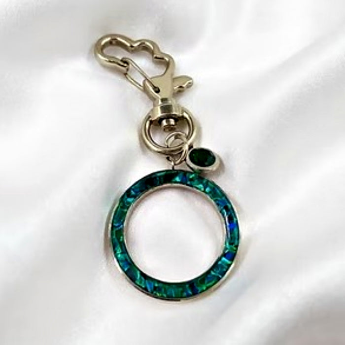 A silver-tone keychain or bag charm featuring a large open circular ring inlaid with iridescent green and blue abalone shell, a small dark green teardrop gemstone, and a heart-shaped clasp, displayed on a white satin fabric background.